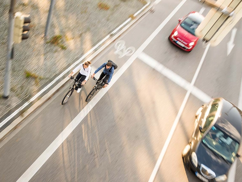 Zwei Fahrradfahrer:innen im Straßenverkehr mit Autos, von schräg vorne/oben fotografiert Zwei Fahrradfahrer:innen im Straßenverkehr mit Autos, von schräg vorne/oben fotografiert