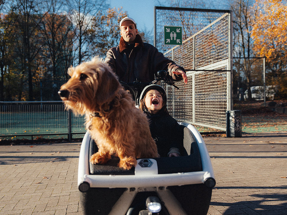 Vater mit Kind und Hund im Lastenrad mit Freudenschrei Vater mit Kind und Hund im Lastenrad mit Freudenschrei